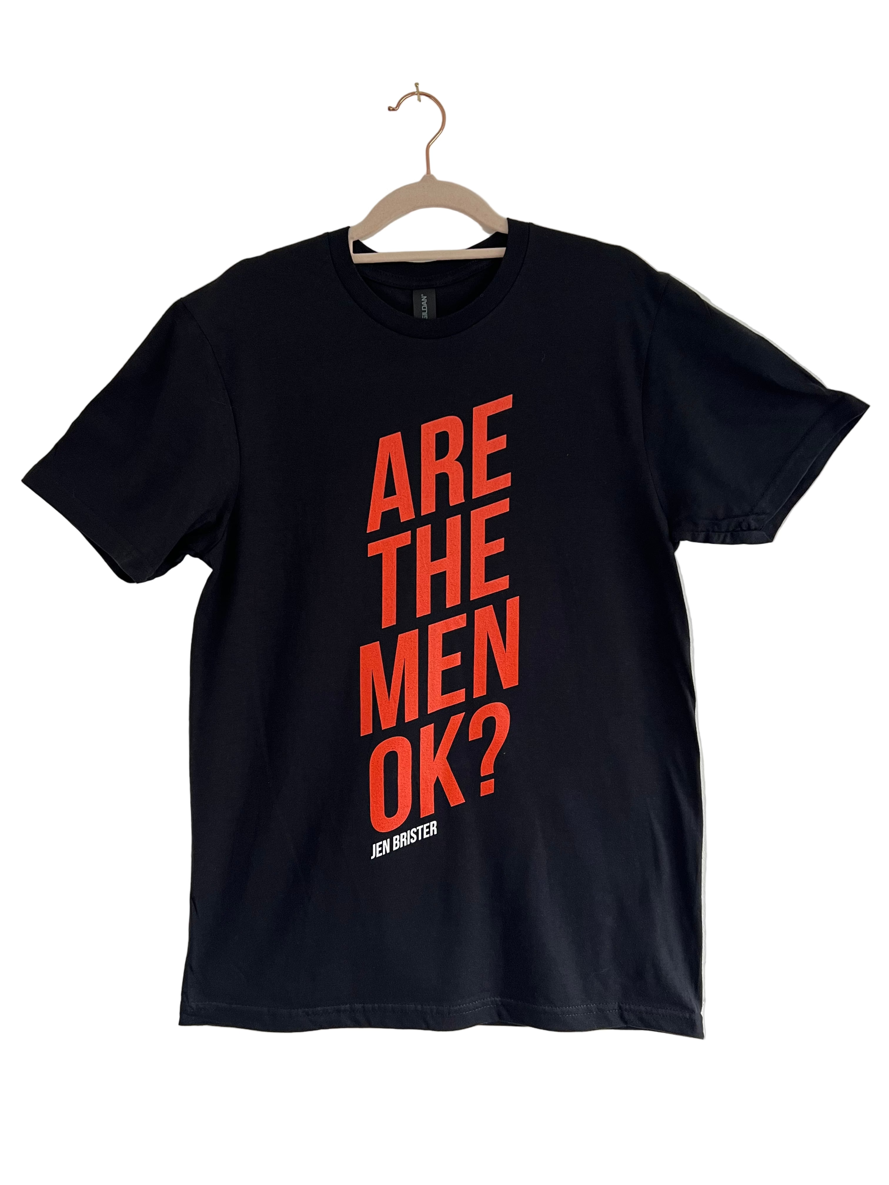 Black t-shirt with orange text 'ARE THE MEN OK?' on a hanger against a white wall.