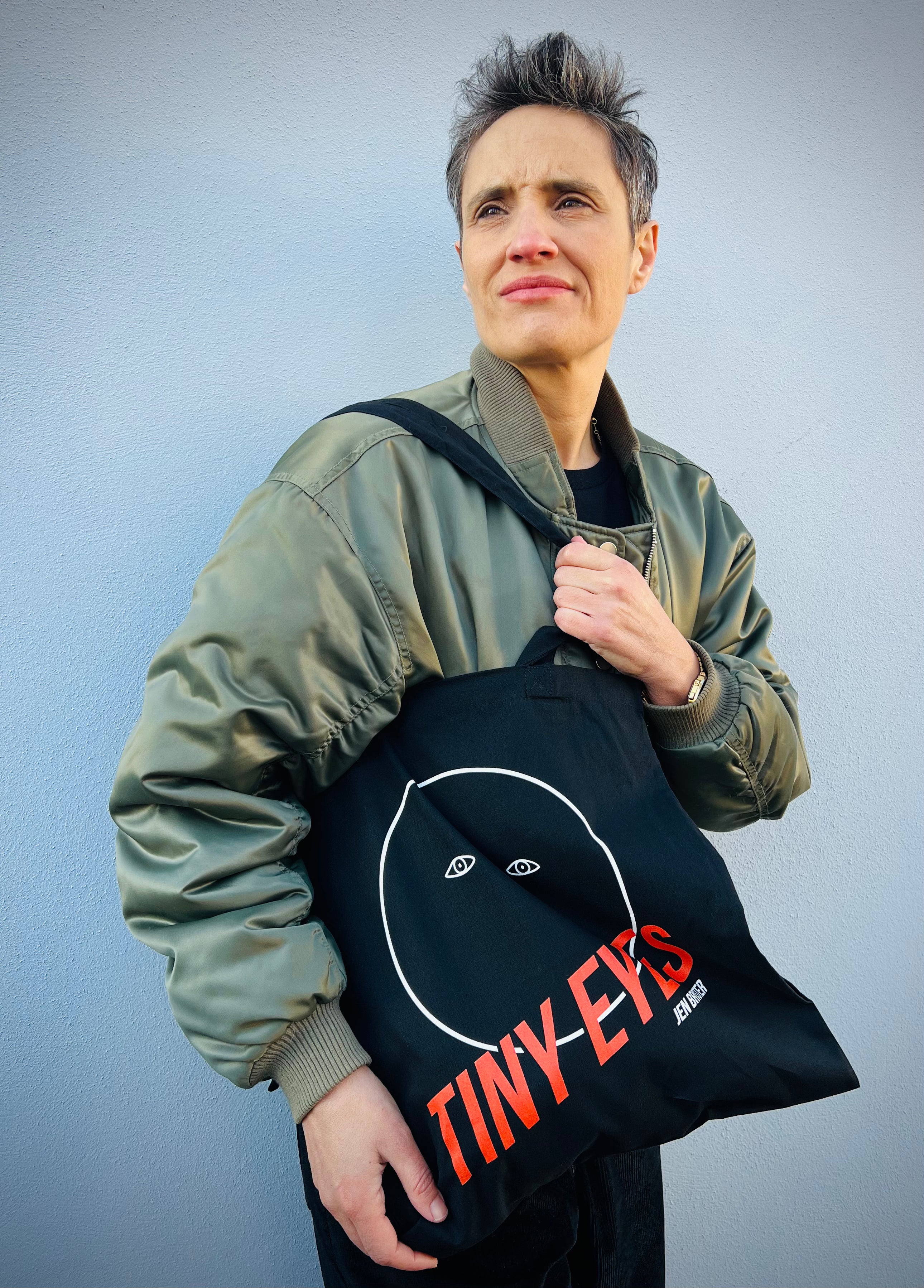 Person holding a black tote bag with 'TINY EYES' branding against a grey background
