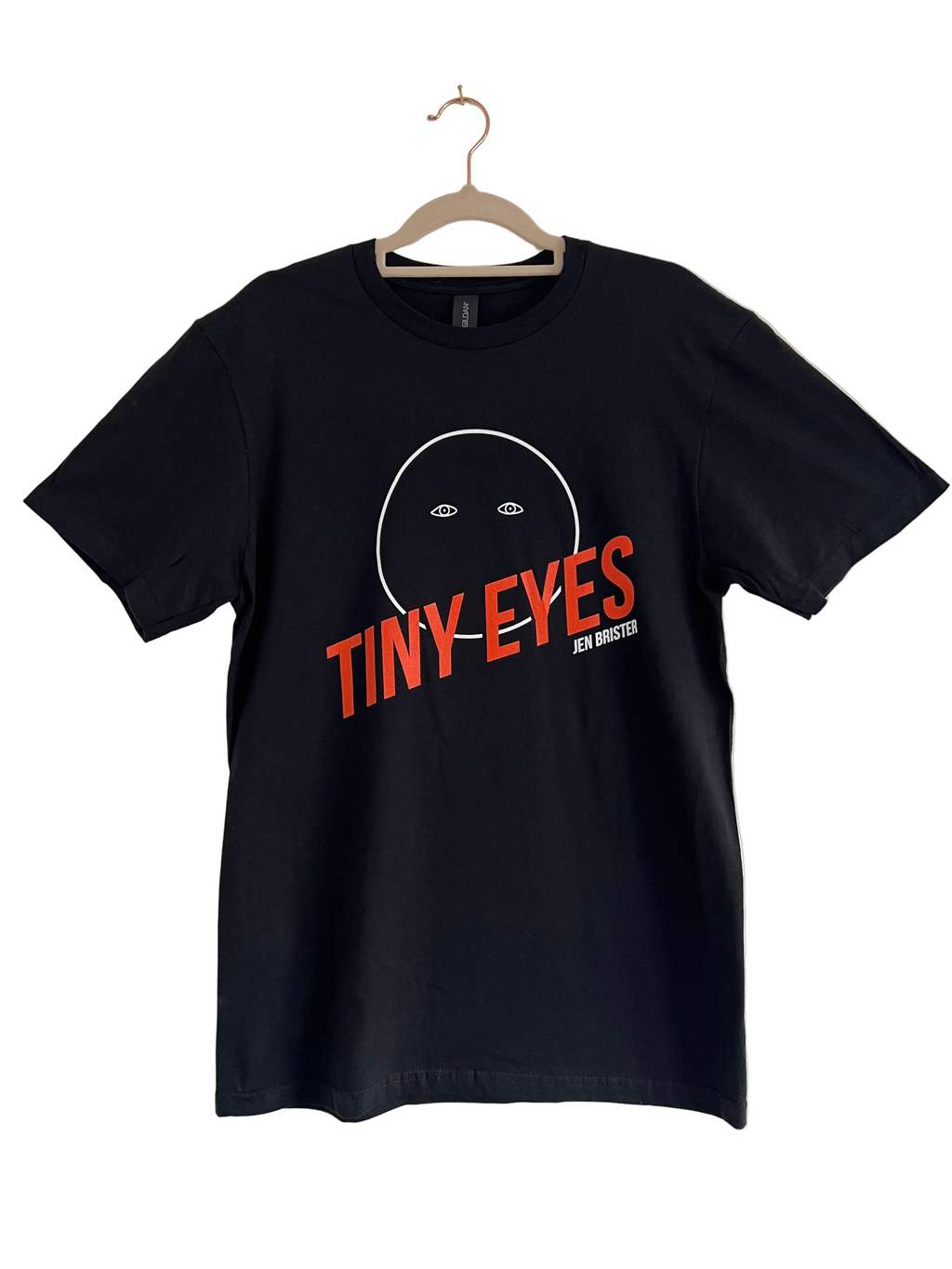 Black t-shirt with 'Tiny Eyes' orange text and graphic on a hanger against a plain background